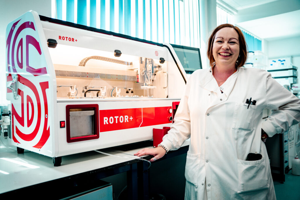 Jane usher  faces the camera and smiles as she stands next to ROTOR+ the high throughput screening robot